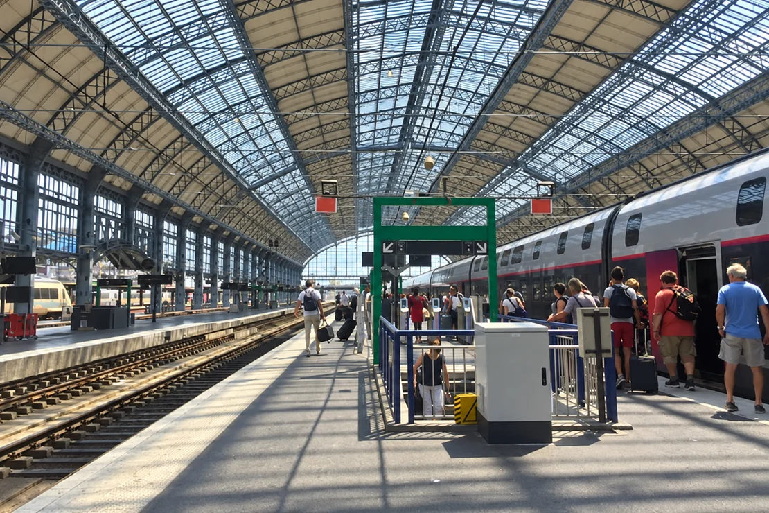 Train à quai et voyageurs sous une grande verrière de gare.